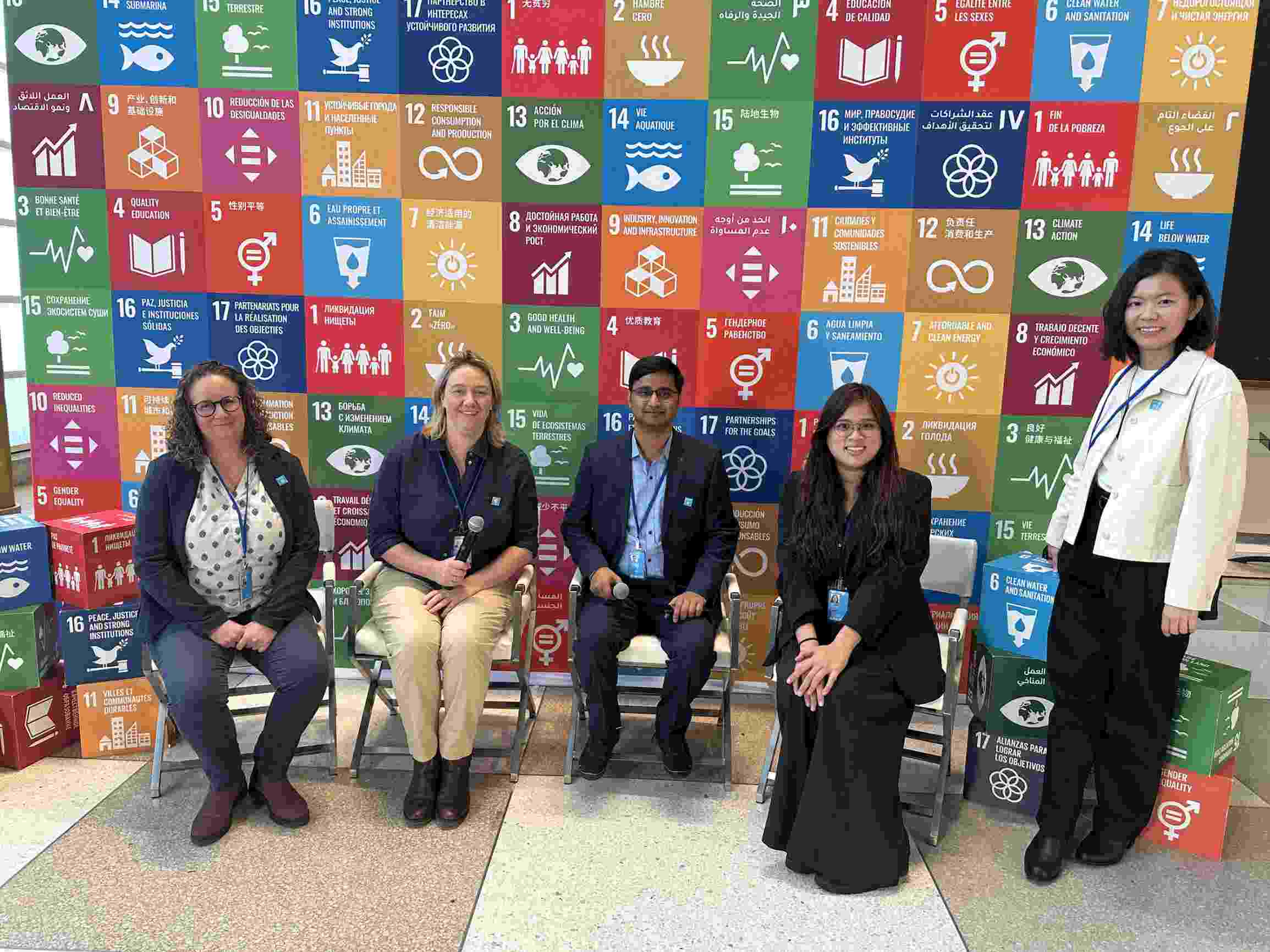 Five Hub members sit and stand together in front of a backdrop of the SDG logos